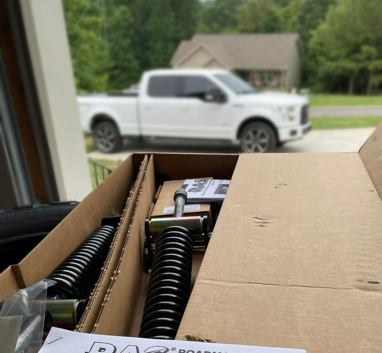 Cardboard box with RoadActive Suspension springs and a label in front of a white truck outside a house.